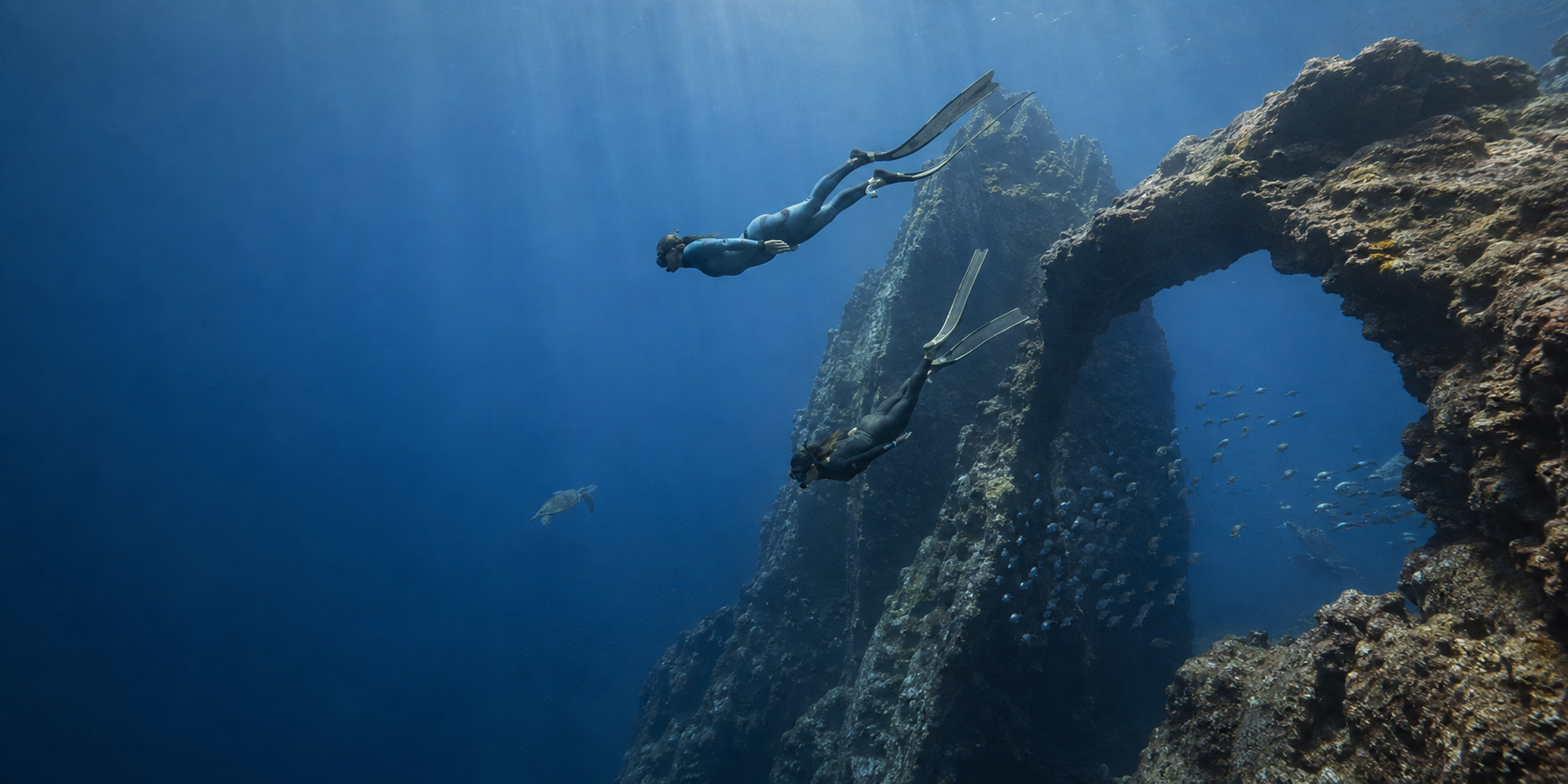 Male and female freedivers in wetsuits and long bi-fins descending beside a natural underwater rock arch, with schooling fish and a sea turtle